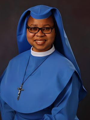 A smiling nun in a blue habit and glasses with a cross necklace.