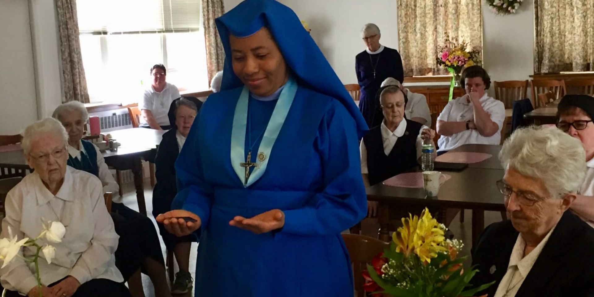 A nun in blue habit reading or praying in a room with others.