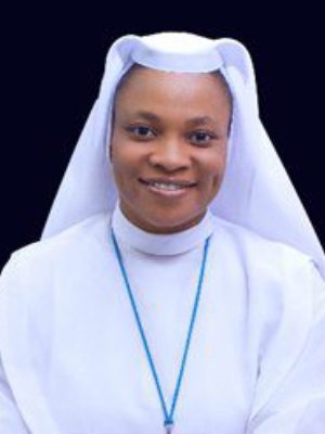 Portrait of a smiling nun in white habit against a black background.