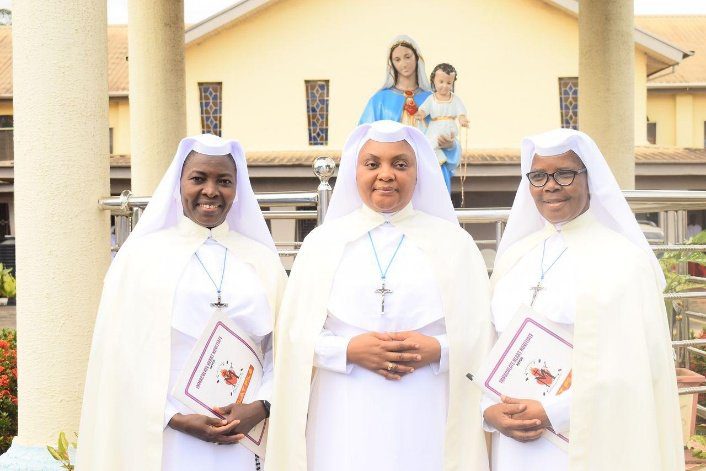 Three nuns smiling and holding books, with two women and a child in the background.