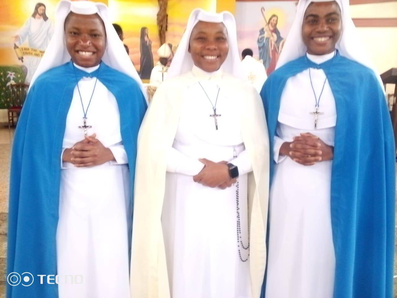 Three smiling nuns in blue and white habits standing together indoors.