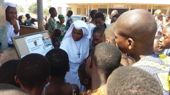 A nun in white habit engaging with local children and adults outdoors.