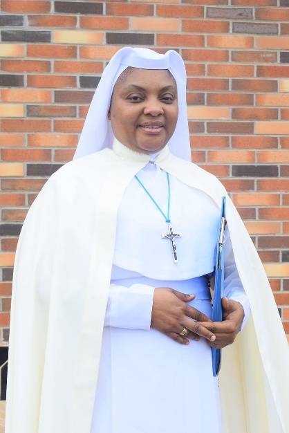 A smiling nun in white habit holding a book, standing against a brick wall.