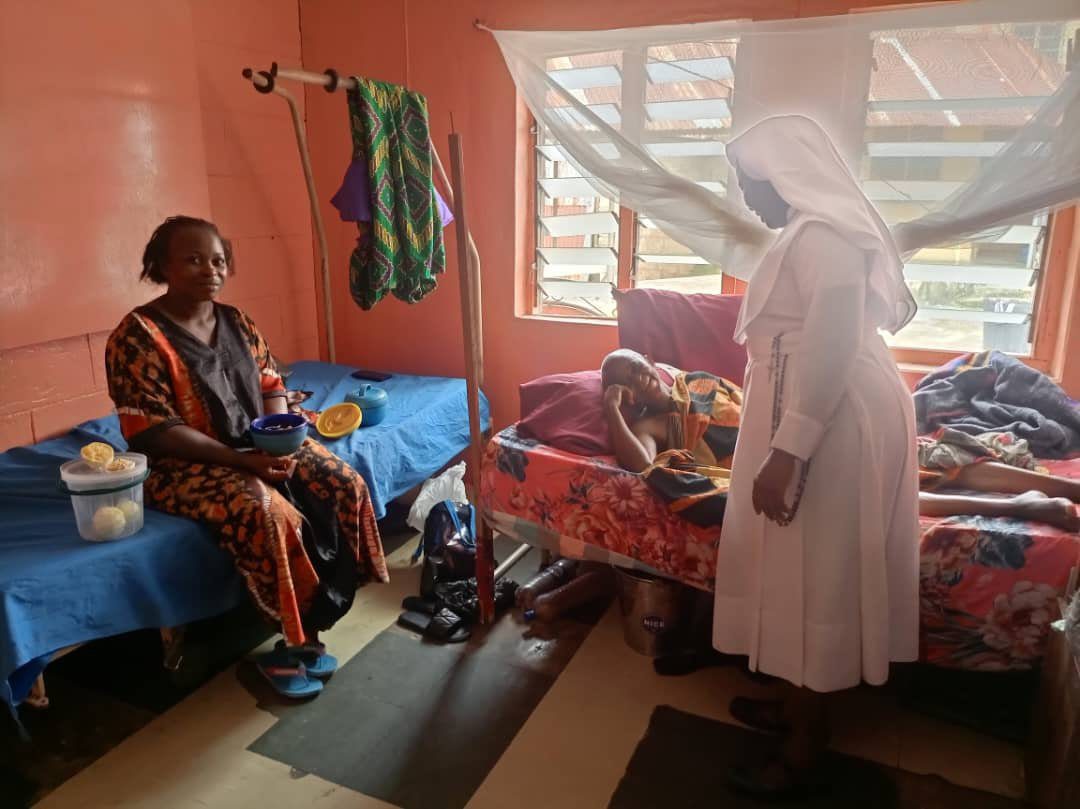 A nurse in white attending to a patient while a woman watches in a modest room.
