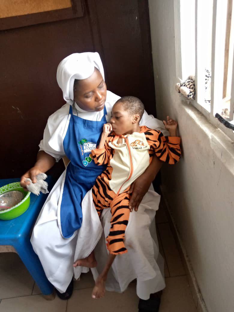 An elderly woman in traditional attire holds a baby in a tiger costume by a window.