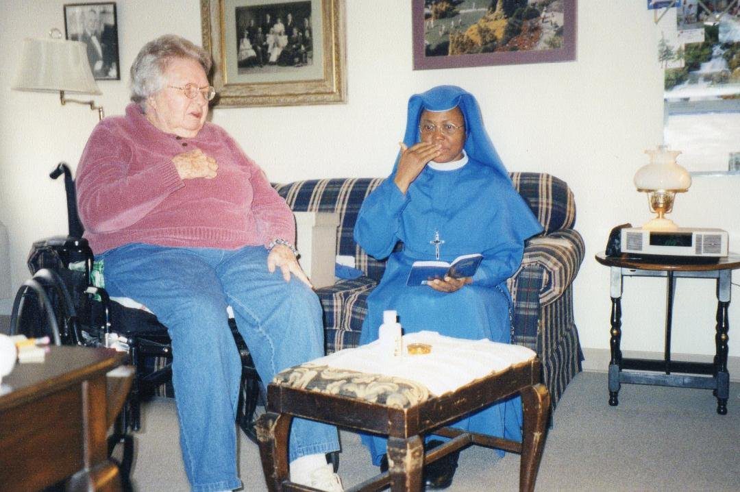 An elderly woman and a nun sitting in a living room with paintings on the wall.