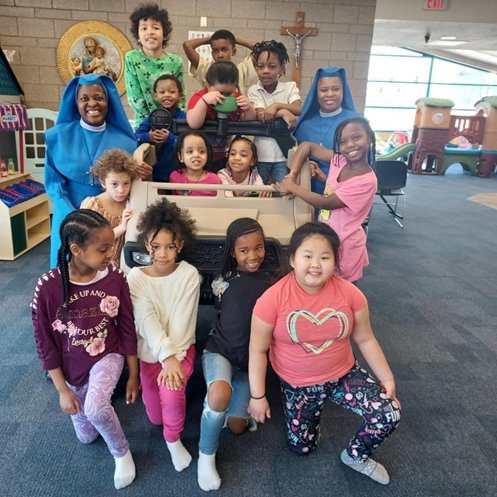 Group of children sitting and standing around a wooden bench indoors.