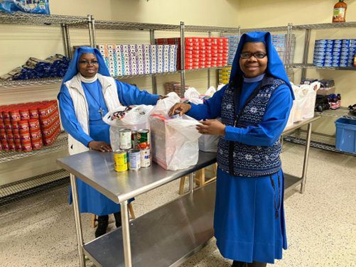 Two women in blue habits packing food items at a table.