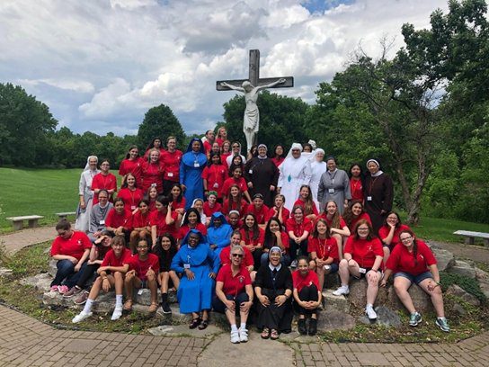 Large group of people in red and blue uniforms posing outdoors by a cross monument.